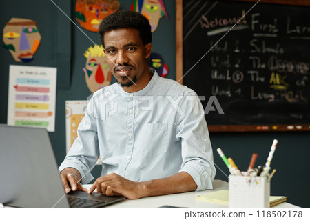 Portrait of smiling African American male teacher captured typing on laptop while sitting at working desk in primary school classroom, copy space Portrait of smiling African American male teacher captured typing on laptop while sitting at working desk in primary school classroom, copy space 118502178