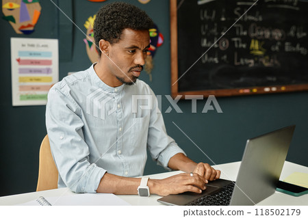 Medium shot of concentrated African American male teacher sitting at working desk typing on laptop in primary school classroom, copy space 118502179