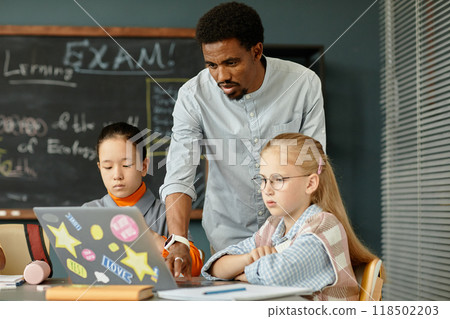African American male teacher helping younger students with using laptop in class typing on computer keyboard in modern classroom at school, copy space 118502203