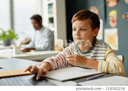 Young concentrated boy keeping finger on touchpad of laptop and looking at screen while studying in sunlit classroom, copy space 118502216