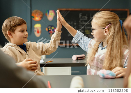 Side view of young blonde schoolgirl sitting at desk happily giving high five to male classmate while having fun with friends during break at school 118502298