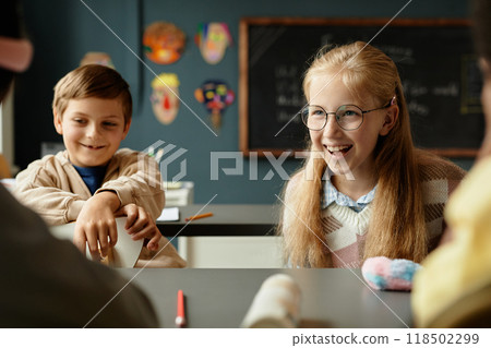 Medium shot of laughing young girl with long blonde hair and glasses sitting at desk having fun with classmates during break at school, copy space Medium shot of laughing young girl with long blonde hair and glasses sitting at desk having fun with classmates during break at school, copy space 118502299