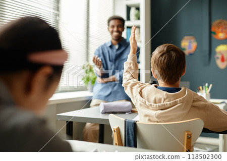 Rear view of young male student sitting at desk raising hand to answer teachers question during lesson in primary school classroom, copy space Rear view of young male student sitting at desk raising hand to answer teachers question during lesson in primary school classroom, copy space 118502300