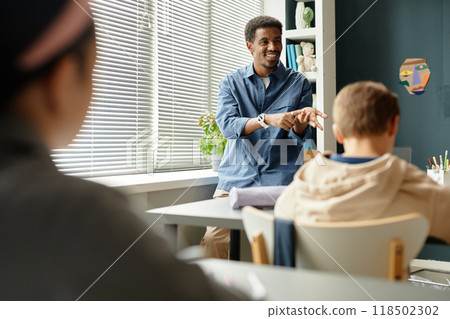 Medium shot of smiling African American male teacher talking to children in classroom with window blinds closed while counting on fingers explaining new topic to students at school, copy space 118502302