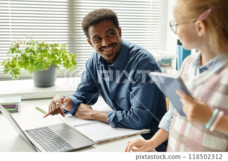 Medium shot of smiling African American male teacher looking at young student while sitting at table with laptop in classroom at primary school, copy space Medium shot of smiling African American male teacher looking at young student while sitting at table with laptop in classroom at primary school, copy space 118502312