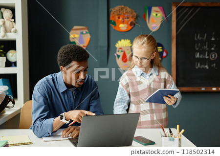 Medium shot of experienced male primary school teacher of African American ethnicity sitting at table talking to young smart student while pointing at laptop screen in classroom, copy space Medium shot of experienced male primary school teacher of African American ethnicity sitting at table talking to young smart student while pointing at laptop screen in classroom, copy space 118502314