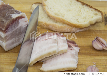 A knife cutting the salo on pieces. Ukrainian products raw lard with peppercorn and garlic, knife, slices of bread, salo lying on wooden board. Ukrainian traditional food concept. A knife cutting the salo on pieces. Ukrainian products raw lard with peppercorn and garlic, knife, slices of bread, salo lying on wooden board. Ukrainian traditional food concept. 118502758
