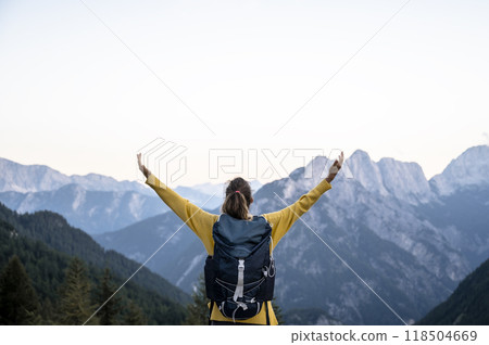 Female hiker with a backpack standing with her arms spread wide looking at a beautiful view of high mountains 118504669