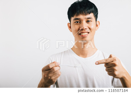A happy Asian young man, in a dental health concept, holds a toothbrush, brushes his teeth, and points to the brush with a smile. Studio shot isolated on white background, presenting oral care. 118507184