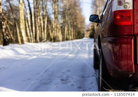 A red car is parked on the snowy side of the road that winds through the forest, and the sun casts long shadows on the snow. A fabulous winter road through the forest. A car on the side of the road. 118507414