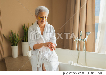 Mature adult old woman applying moisturizing body lotion on hands. Senior lady rubbing nourishing anti age cream doing morning daily skincare routine in bathroom. Beauty treatment spa body skin care Mature adult old woman applying moisturizing body lotion on hands. Senior lady rubbing nourishing anti age cream doing morning daily skincare routine in bathroom. Beauty treatment spa body skin care 118508506