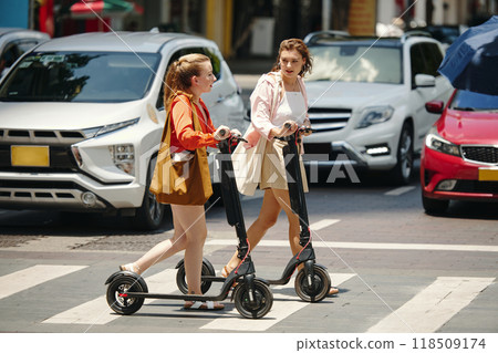 Two Women Riding Electric Scooters on City Street 118509174