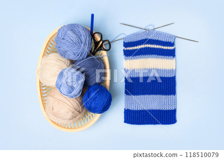 Blue and beige knitting balls in basket with knitting needles and knitted scarf on blue background. Set for the hobby of knitting warm clothes or toys. Concept needlework 118510079