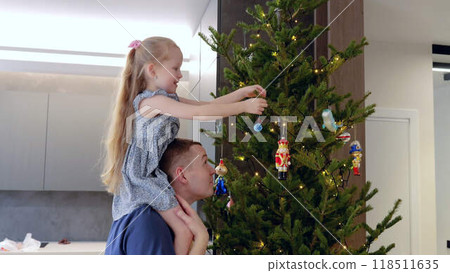 Touching moment father helps daughter place decorations on Christmas tree. Father and daughter work together to decorate Christmas tree with daughter perched on his shoulders winter holidays. 118511635