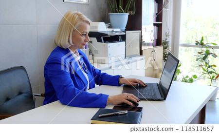 Caucasian business woman in suit sits at office desk working on laptop office job successful career. In office setting mid adult businesswoman in suit focuses on computer work professional success. Caucasian business woman in suit sits at office desk working on laptop office job successful career. In office setting mid adult businesswoman in suit focuses on computer work professional success. 118511657