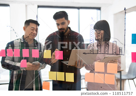 Diverse team brainstorming with sticky notes and graphs in office Diverse team brainstorming with sticky notes and graphs in office 118511757