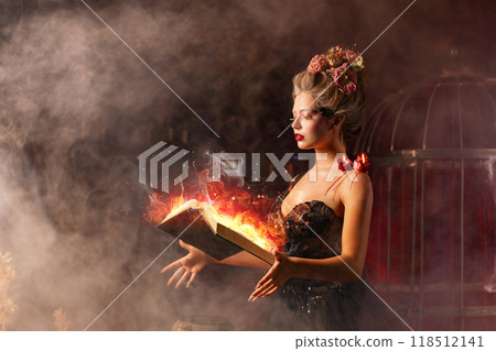 Young witch with glowing book of spell in dark 118512141