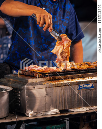 Person Grilling Meat With Tongs at a Food Stall Person Grilling Meat With Tongs at a Food Stall 118513291