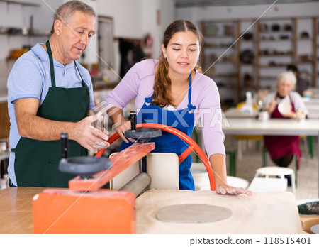Elderly potter teaches woman how to roll clay on a craft machine clay press roller Elderly potter teaches woman how to roll clay on a craft machine clay press roller 118515401