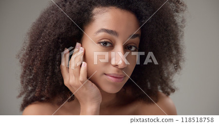 Beauty and healthcare concept - beautiful African American woman with curly afro hairstyle and clean, healthy skin touches her cheek and face with her hand, posing and looking at the camera 118518758