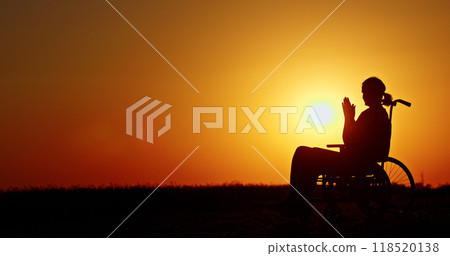 Silhouette of a person in a wheelchair at sunset, hands clasped in quiet reflection. 118520138