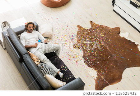 Young man relaxing on sofa with confetti chaos on floor in living room. Top view of home interior after celebration party. 118525432