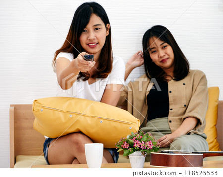 Two women relaxing on couch, one holding TV remote and other resting with content smile, with ukulele and potted plant nearby 118525533