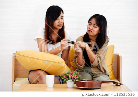 Two women on couch playfully passing TV remote, with one woman showing reluctant expression. ukulele and potted plant rest on table 118525536