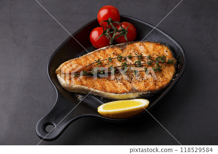Grilled salmon with cherry tomatoes, thyme and lemon on a small black grill pan. Fried fish is a traditional dish of home cooking and restaurants. Black background, selective focus, close-up. 118529584