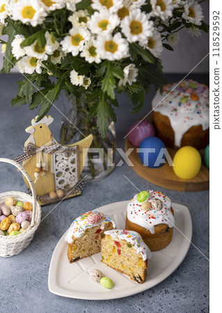 Two small Easter kulichs with candied fruits in white glaze with colorful sprinkles in the cut. Painted chicken and quail eggs. Traditional Easter baking. Easter holiday. Close-up, selective focus. Two small Easter kulichs with candied fruits in white glaze with colorful sprinkles in the cut. Painted chicken and quail eggs. Traditional Easter baking. Easter holiday. Close-up, selective focus. 118529592