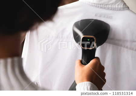 Over the shoulder angle of a young Caucasian woman smoothing a white man's shirt with a handheld black steamer on a hanger 118531314
