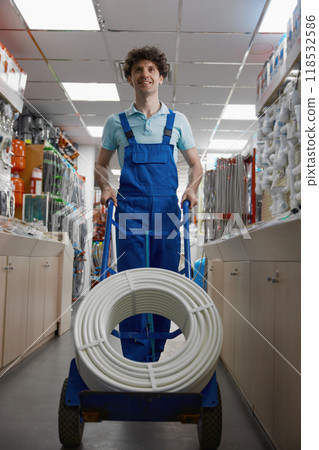 Satisfied plumber in coveralls pushing trolley cart with purchased water hose 118532586