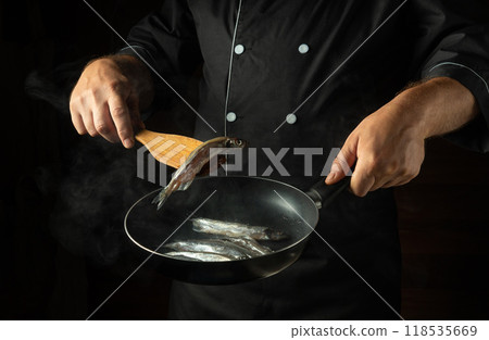 Cooking fish in a frying pan. A cook fries capelin in a restaurant kitchen before dinner. A spatula with a small fish in the chef hand 118535669
