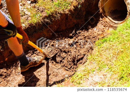 On construction site, worker digs trench with shovel in order to install drainage pipe 118535674