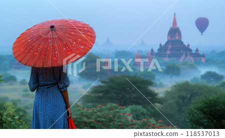 Burmese Woman Holding Traditional Red Umbrella Gazes At Hot Air Balloon Over Bagan Plain On Misty Morning In Mandalay, Myanmar. Burmese Woman Holding Traditional Red Umbrella Gazes At Hot Air Balloon Over Bagan Plain On Misty Morning In Mandalay, Myanmar. 118537013