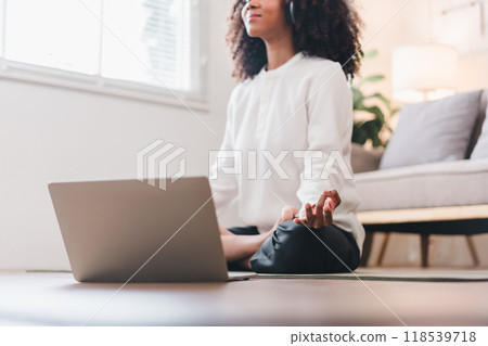 Relaxed African American woman meditating in seated position on yoga mat at home, listening to music with headphones. 118539718