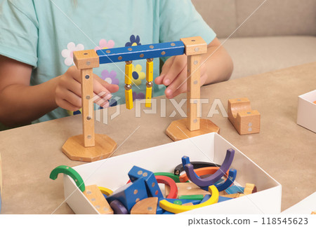 Girl assembles swing from magnetic wooden construction set. Toys and activiti children. Developmental activities at home. Girl assembles swing from magnetic wooden construction set. Toys and activiti children. Developmental activities at home. 118545623