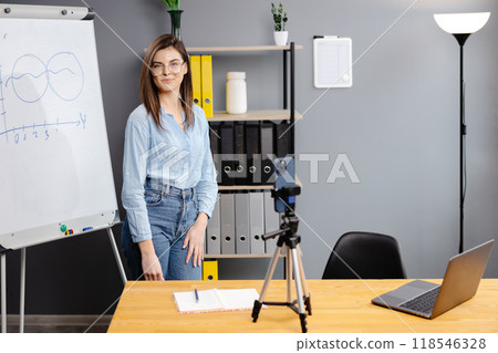 Positive blogger woman speaking at smartphone webcam on tripod, recording post for vlog. Online teacher, coach working, giving webinar at home. Presenter taking video on cellphone 118546328