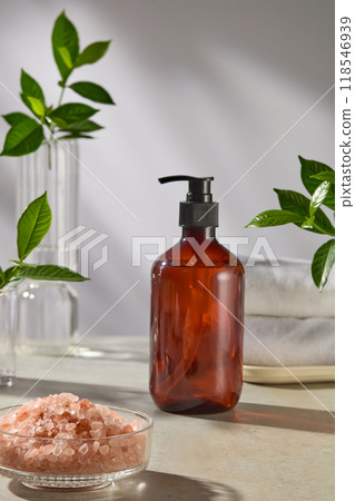 Cosmetic advertising pic in spa space background, an amber pump bottle without label displayed in center, next to a glass bowl of pink salt and glass jar. Green plants decorated in flanked 118546939