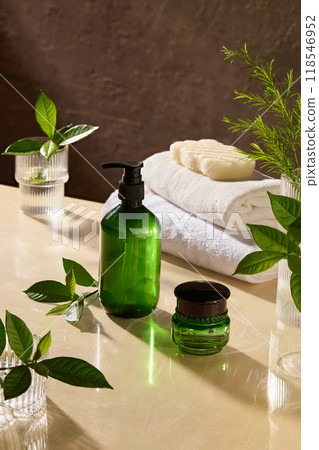 Natural spa theme photo with unlabeled green pump bottle and moisturizer pot placed by side on light yellow spa table top, next to some glass jars contain green plant, bath towel and soap Natural spa theme photo with unlabeled green pump bottle and moisturizer pot placed by side on light yellow spa table top, next to some glass jars contain green plant, bath towel and soap 118546952