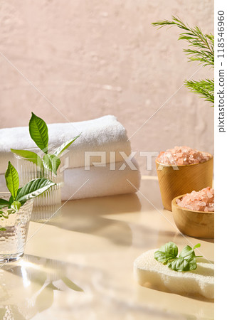 Creative photo for advertising in spa space design with lots of sunlight. Beige table displaying various spa things like spa pink salt, bath towels, sponge, glass jars, green plants. Displaying space 118546960