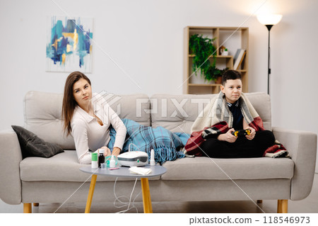 Young woman holding a mask nebulizer and inhaling fumes medication into lungs. Sick lady inhaling through inhaler mask at home and funny boy with gamepad playing video game at home near her Young woman holding a mask nebulizer and inhaling fumes medication into lungs. Sick lady inhaling through inhaler mask at home and funny boy with gamepad playing video game at home near her 118546973