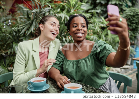High angle shot of biracial best friends taking selfie while drinking hot black tea in eco cafe High angle shot of biracial best friends taking selfie while drinking hot black tea in eco cafe 118554848