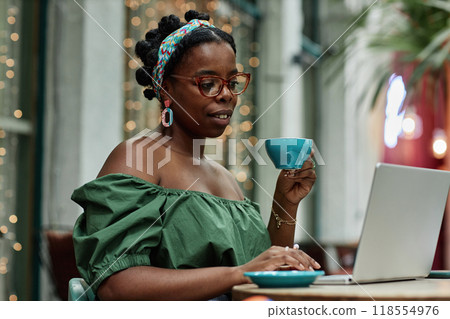Young African American woman holding cup of tea while surfing in Internet in cafe Young African American woman holding cup of tea while surfing in Internet in cafe 118554976