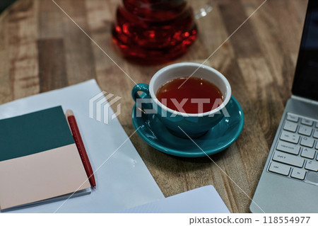High angle shot of turquoise ceramic cup of black tea on wooden table with laptop and notes High angle shot of turquoise ceramic cup of black tea on wooden table with laptop and notes 118554977