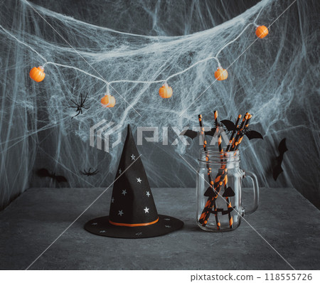 Halloween beverage table setting with glass jar, drinking straws, witch hat and cobwebs on dark gray background. Halloween beverage table setting with glass jar, drinking straws, witch hat and cobwebs on dark gray background. 118555726