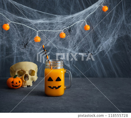 Halloween pumpkin or carrot drink in a glass jar with jack lantern face. Halloween party decorations with juice, skull and cobwebs on blue background. Copy space. 118555728