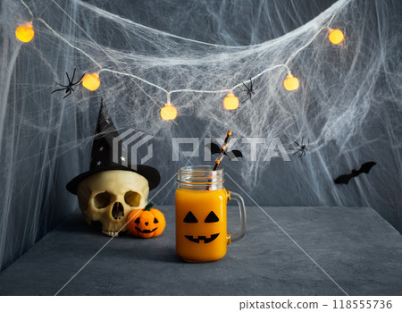 Halloween drink on blue background with copy space. Pumpkin juice in a glass jar with a creepy smile, skull and glowing garland. Halloween drink on blue background with copy space. Pumpkin juice in a glass jar with a creepy smile, skull and glowing garland. 118555736