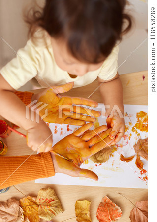 little child girl and mother painting hands and autumn leaves together. mom and baby showing hands in yellow paint. top view little child girl and mother painting hands and autumn leaves together. mom and baby showing hands in yellow paint. top view 118558289