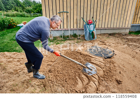 Builder compacts sand near septic tank manhole cover, patting its surface with shovel. 118559408